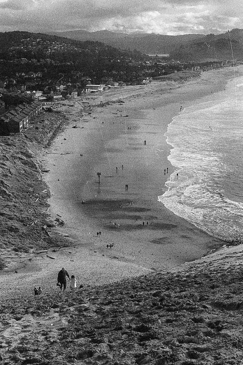 A high-angle view shows a wide beach with waves breaking on the shore, a town along the coast, and mountains in the distance.