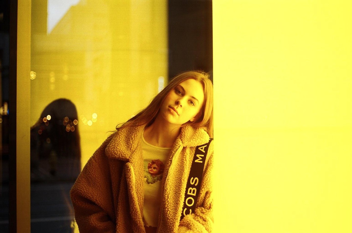 A woman with long hair wears a teddy coat and a t-shirt with a cherub print, leaning against a bright yellow wall with a reflection of another person in the background.