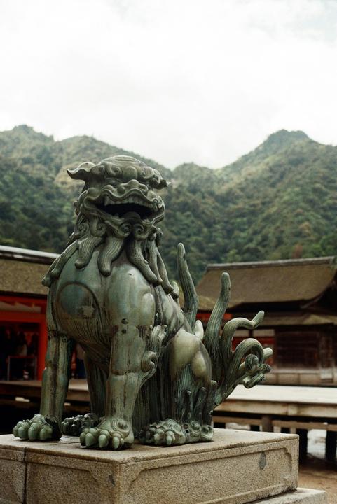 A stone lion statue sits on a pedestal with mountains and buildings in the background.