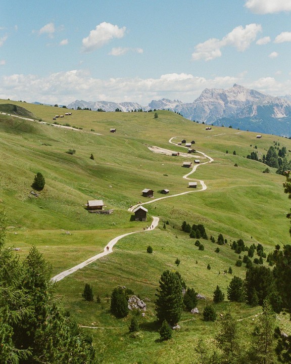 A winding path leads through a green valley dotted with small wooden huts, with mountains in the background.