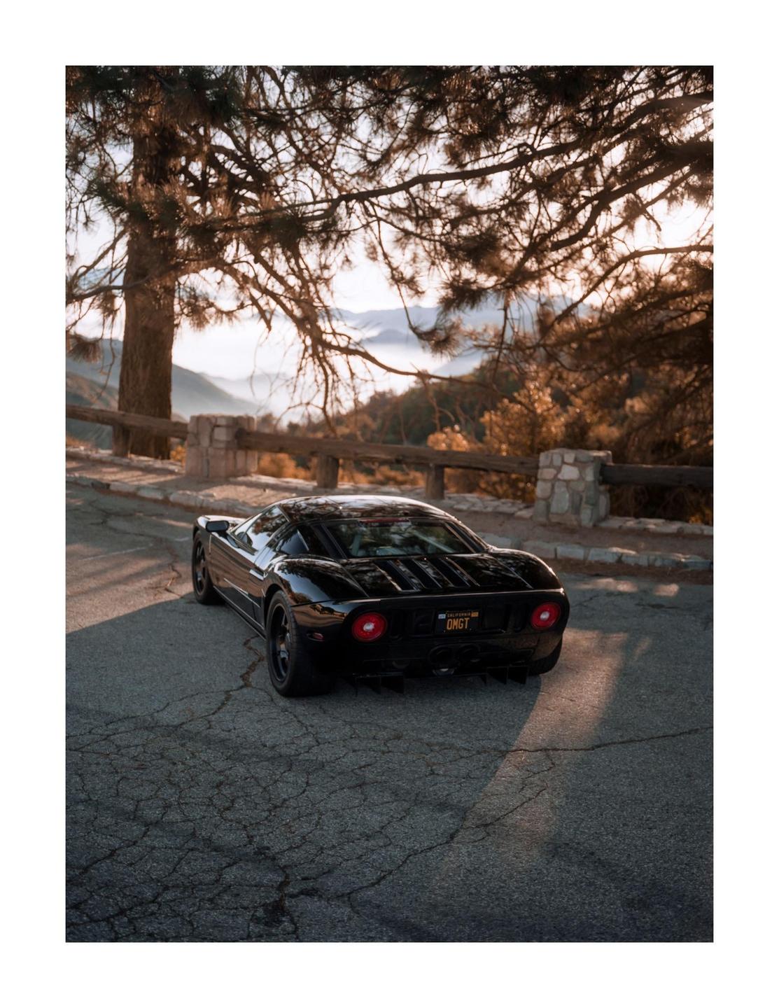 A black sports car is parked on a cracked asphalt road with a mountain landscape in the background.