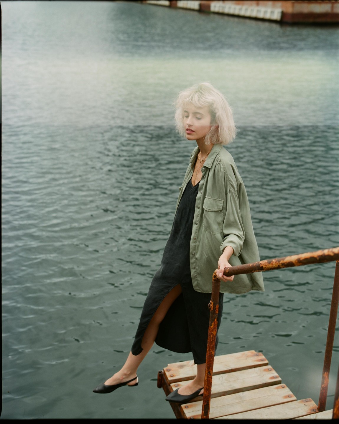 A woman with blonde hair stands on a small wooden dock next to water, holding onto a rusty railing.