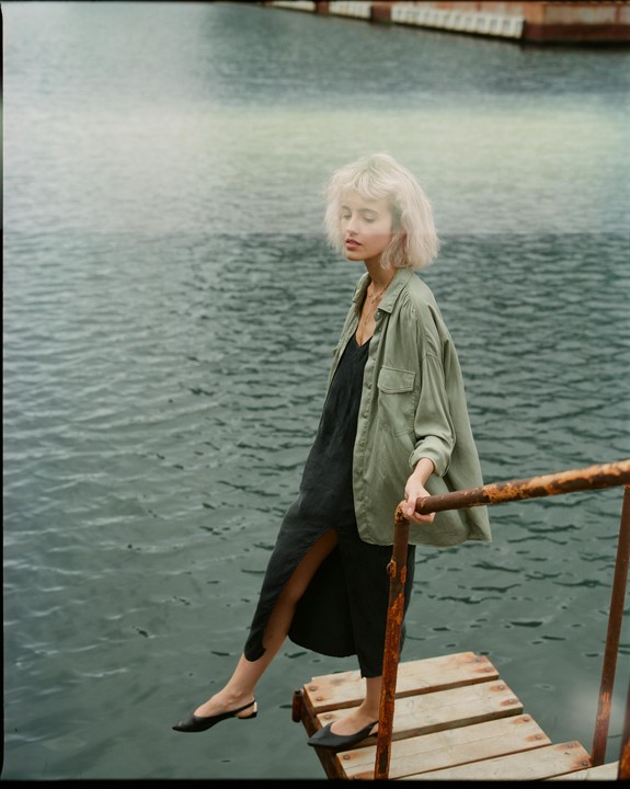 A woman with blonde hair stands on a small wooden dock next to water, holding onto a rusty railing.