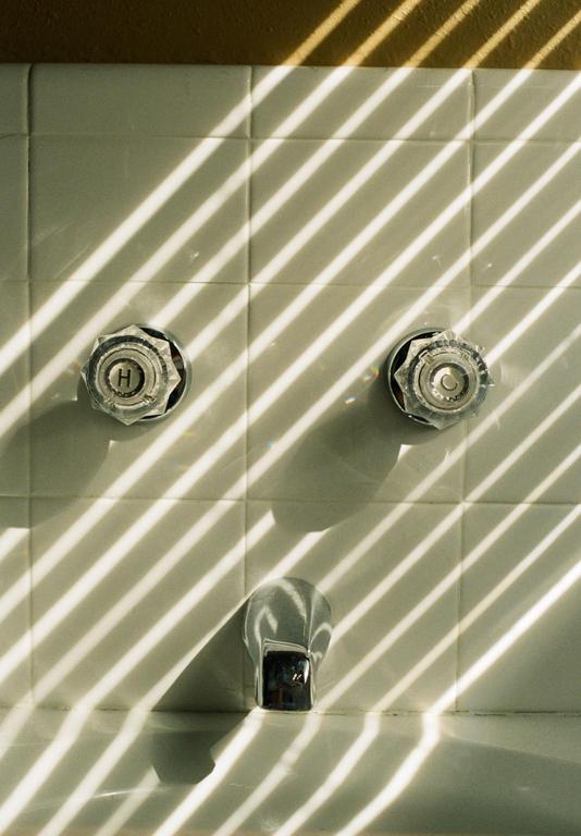 Sunlight streams through a window, casting striped shadows across a tiled bathroom wall with two faucet handles and a spout.