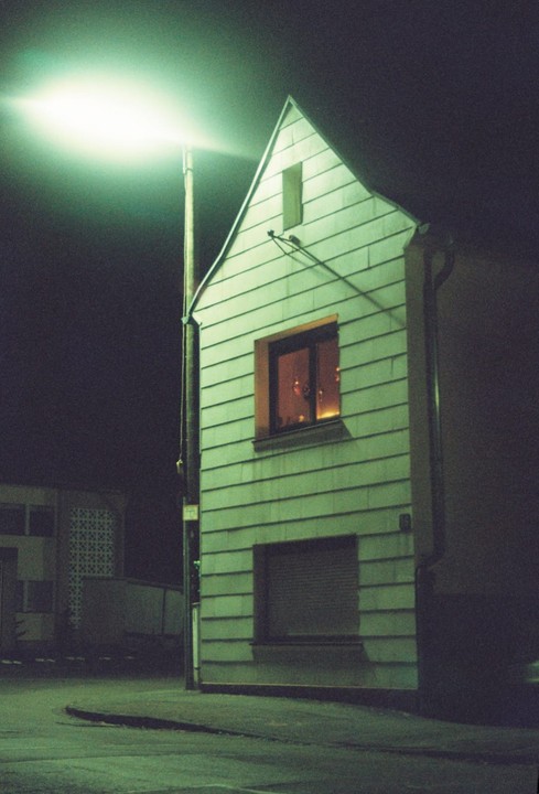 A narrow, light green house with a glowing window stands next to a bright streetlamp at night.