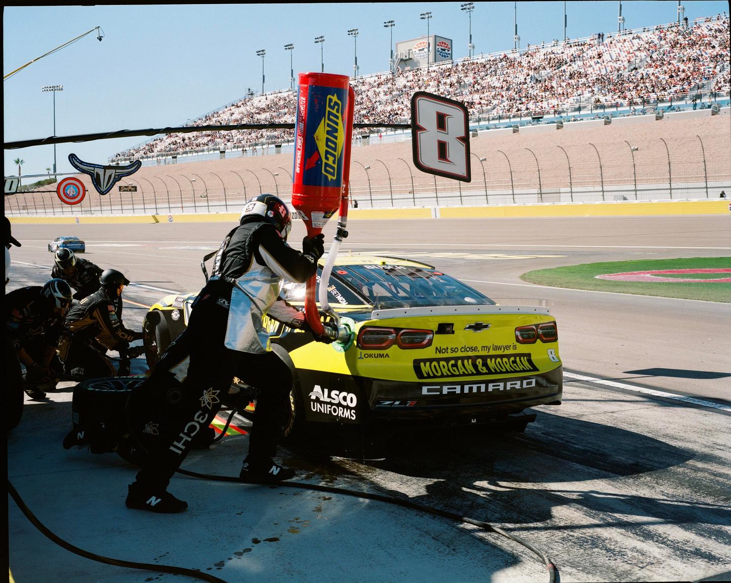 A pit crew refuels a yellow race car at a speedway while spectators watch from the stands.