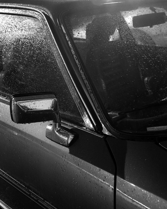 Raindrops cover the side window, windshield, and body of a car.