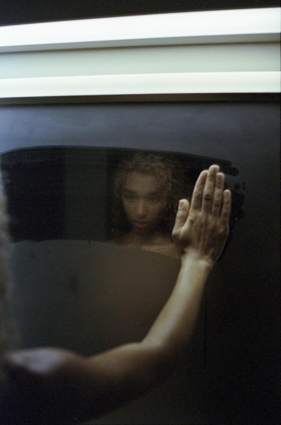A woman with curly hair looks at her reflection in a dark surface, with her hand pressed against it.