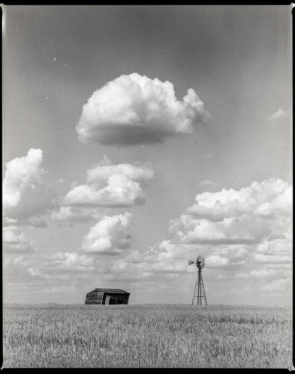A small wooden shack and a windmill stand in a field under a cloudy sky.