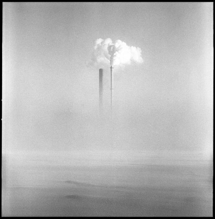 Two industrial smokestacks emit plumes of smoke into a hazy sky above a flat, obscured landscape.