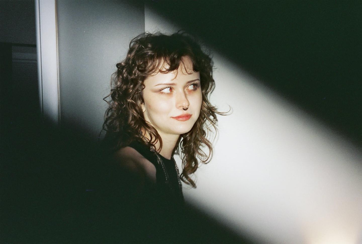 A young woman with curly hair and a nose ring looks to the side, illuminated by a diagonal beam of light.