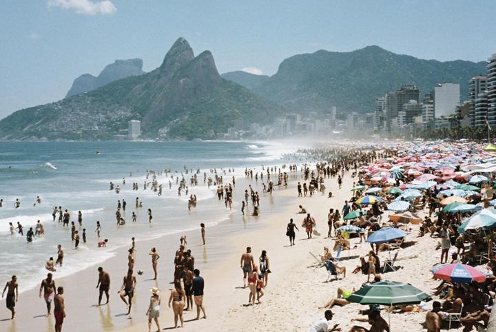 A crowded beach with many people in the water and under colorful umbrellas, with mountains and a city in the background.