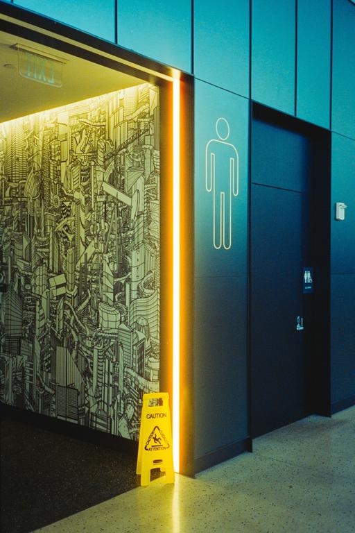 A yellow caution sign stands on the floor next to a wall with a city mural and a men's restroom sign.