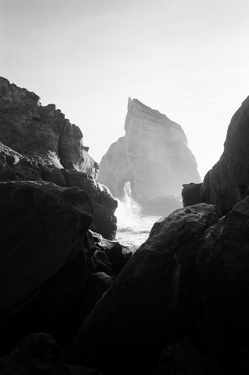 A large rock formation rises from the ocean as waves crash against it, viewed from a rocky shore.