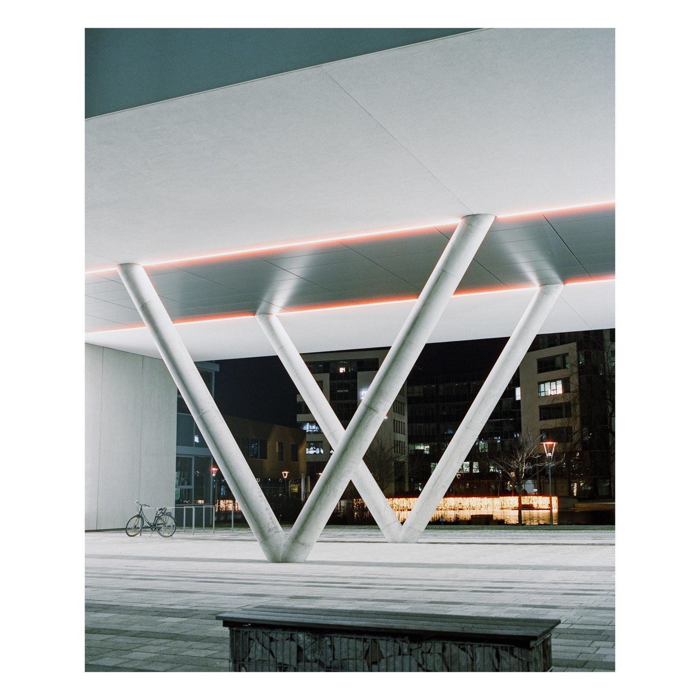 A bicycle is parked under a modern building with angled support columns and a neon light strip at night.