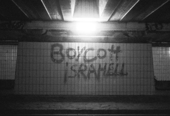 Graffiti that reads "Boycott Israhell" is visible on a tiled wall in a dark underpass.