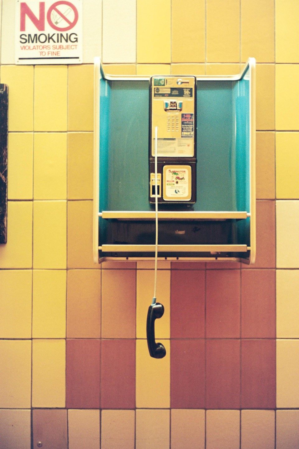 A payphone receiver hangs from its cord against a tiled wall with a "No Smoking" sign.