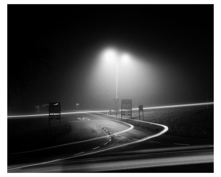 A winding road with light trails is illuminated by a street light on a foggy night.