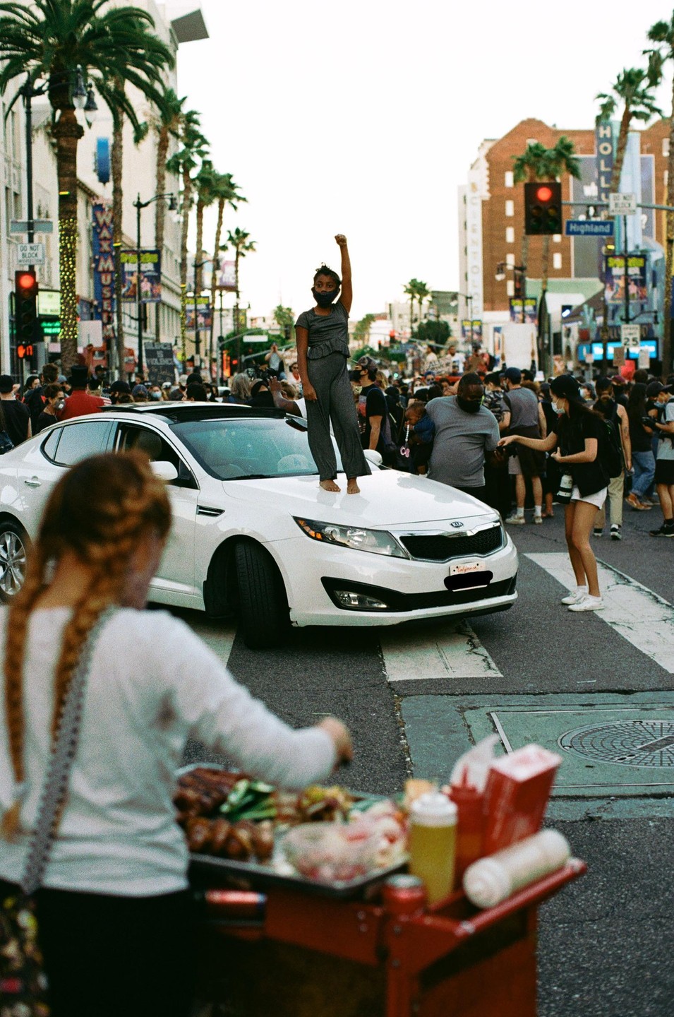A child stands on the hood of a white car with a raised fist in the middle of a street lined with palm trees and buildings.