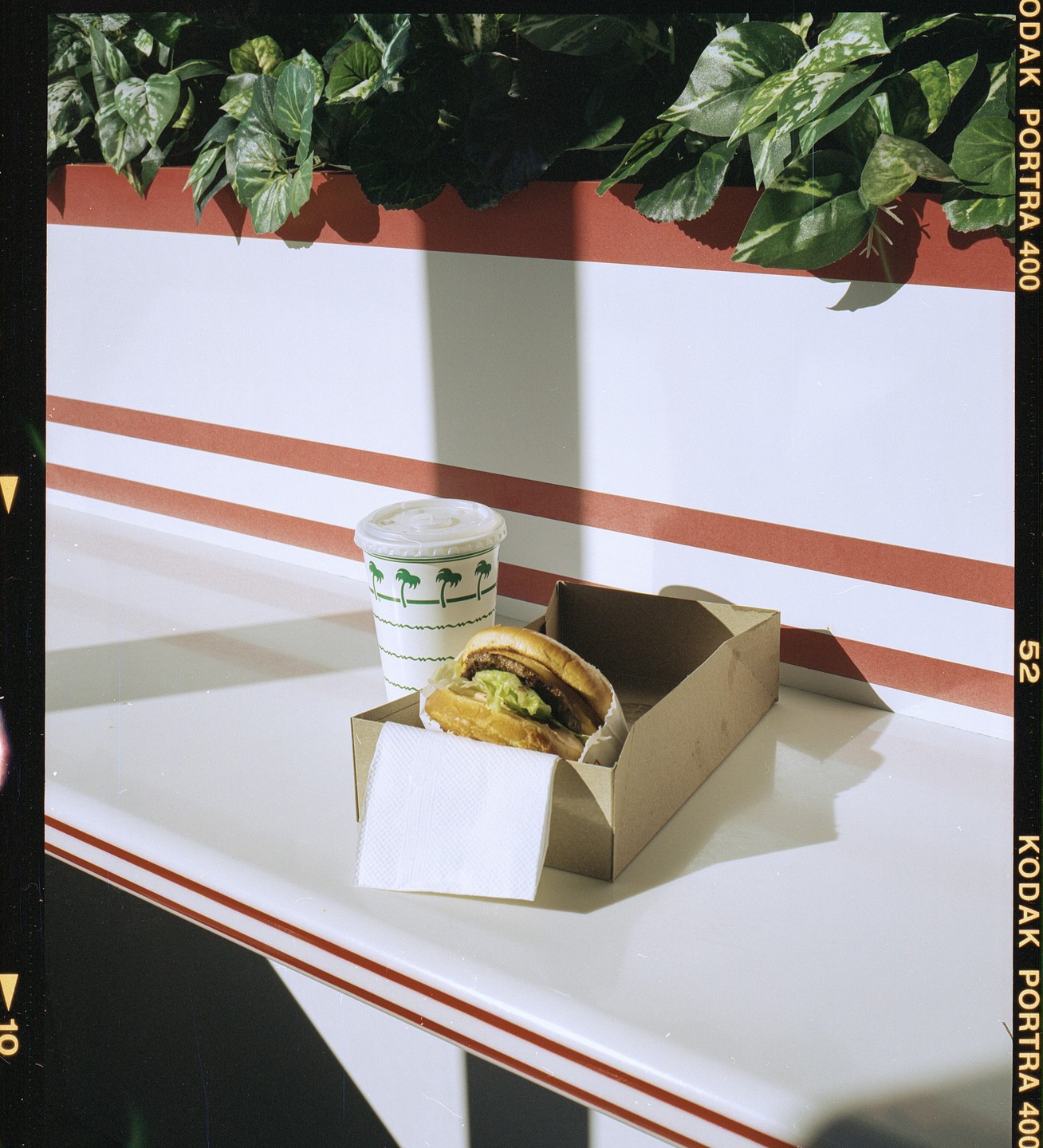A burger in a cardboard box with a napkin and a drink cup sit on a white counter with red stripes, beneath green foliage.