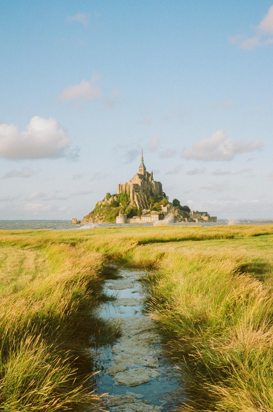A medieval abbey stands on a rocky island surrounded by water and a vast expanse of golden marsh grass.