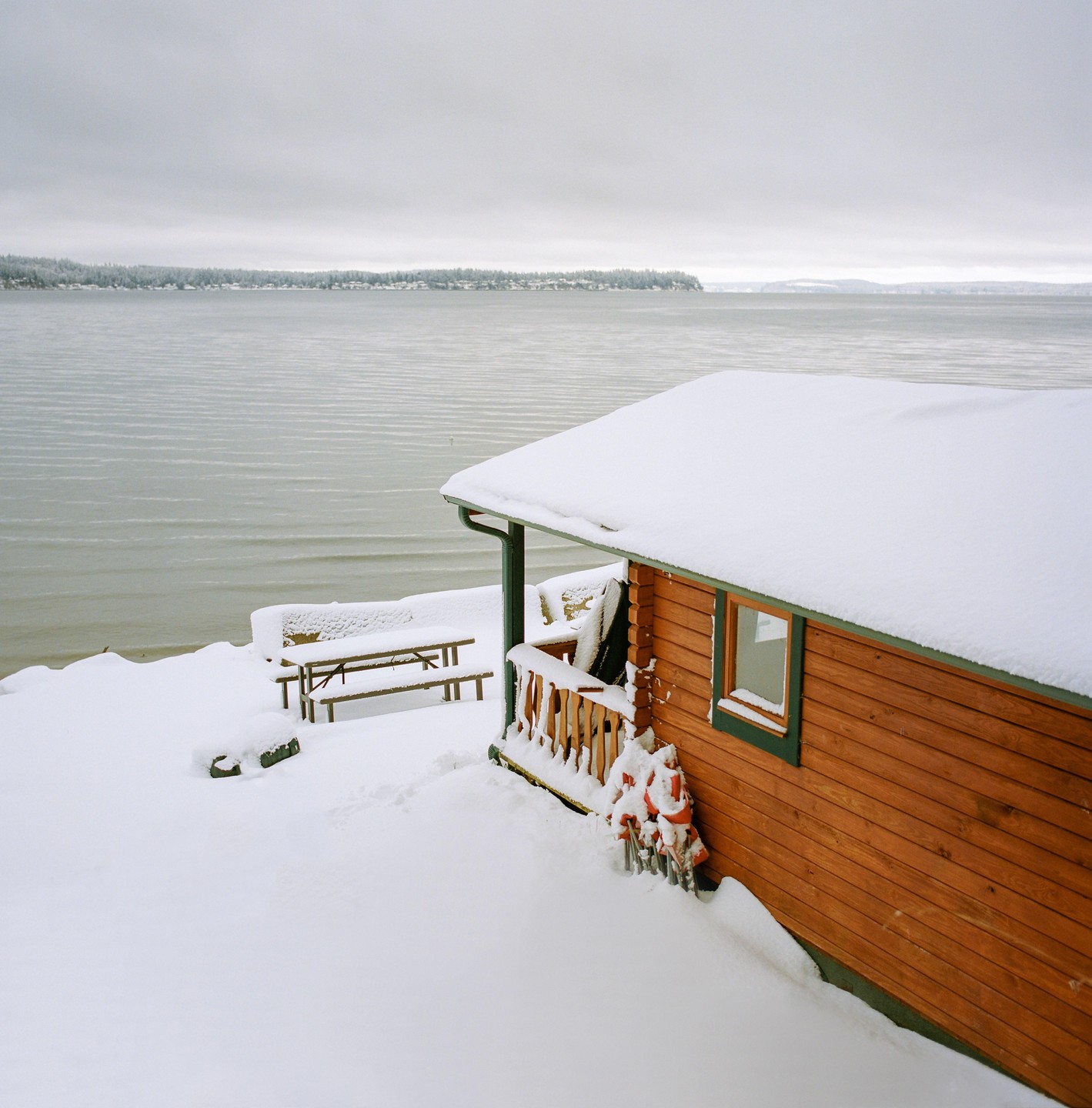 A wooden cabin with a snow-covered roof sits beside a body of water with a distant shoreline.