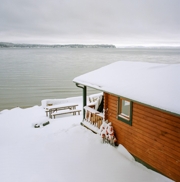 A wooden cabin with a snow-covered roof sits beside a body of water with a distant shoreline.