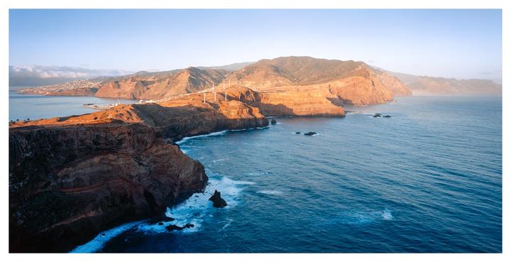A panoramic landscape shows a rugged coastline with cliffs meeting the ocean, a town in the distance, and wind turbines on a hill.