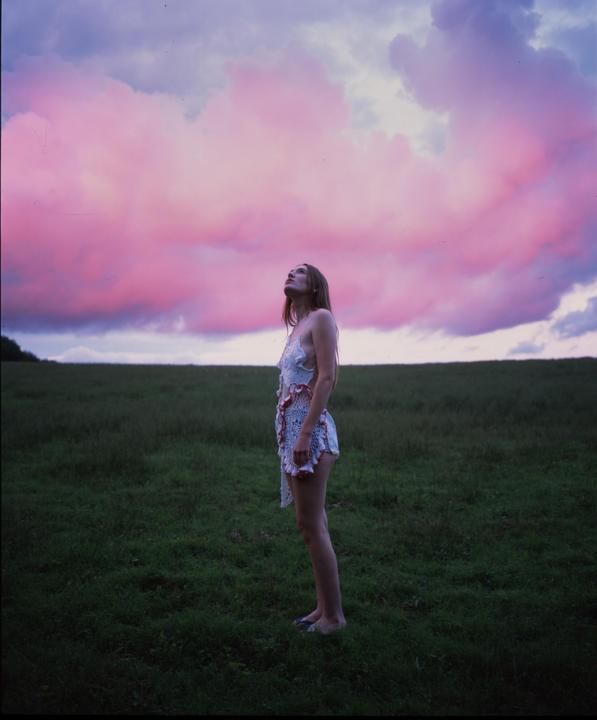 A woman stands in a field looking up at a sky filled with pink clouds.