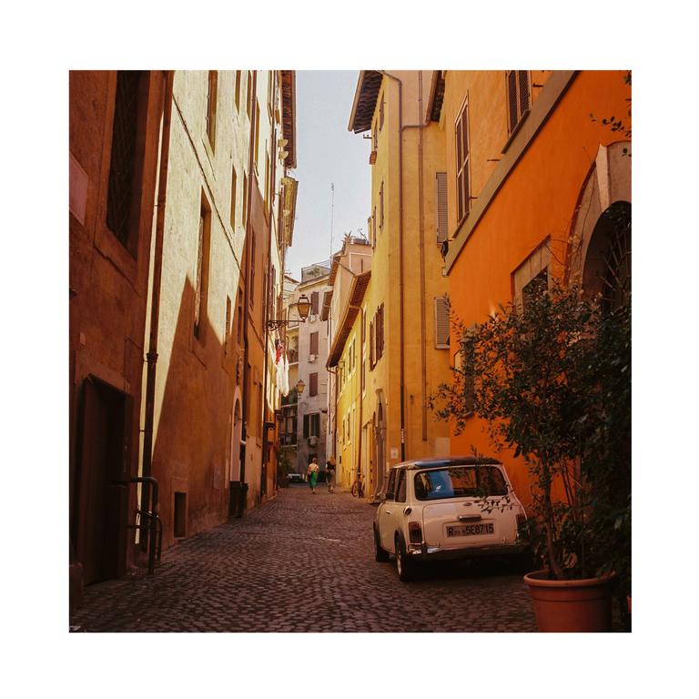 A white car is parked on a cobblestone street between tall buildings with a person walking in the distance.