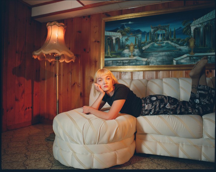 A woman with blonde hair lies on a sofa in a wood-paneled room with a large painting and a floor lamp.