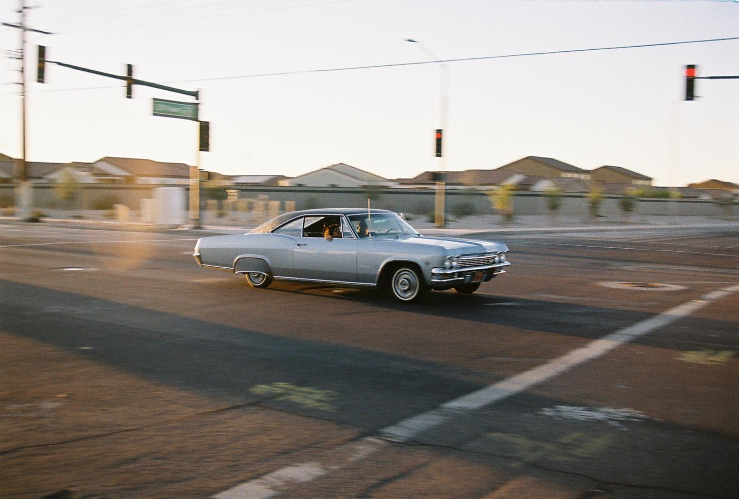 A silver vintage car with a dog in the passenger seat drives down a street at sunset.