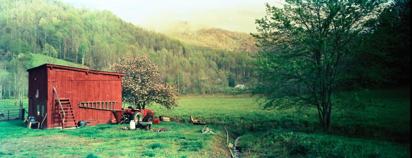 A red barn stands in a green field with a blooming tree and a tractor, with a forest and mountains in the background.