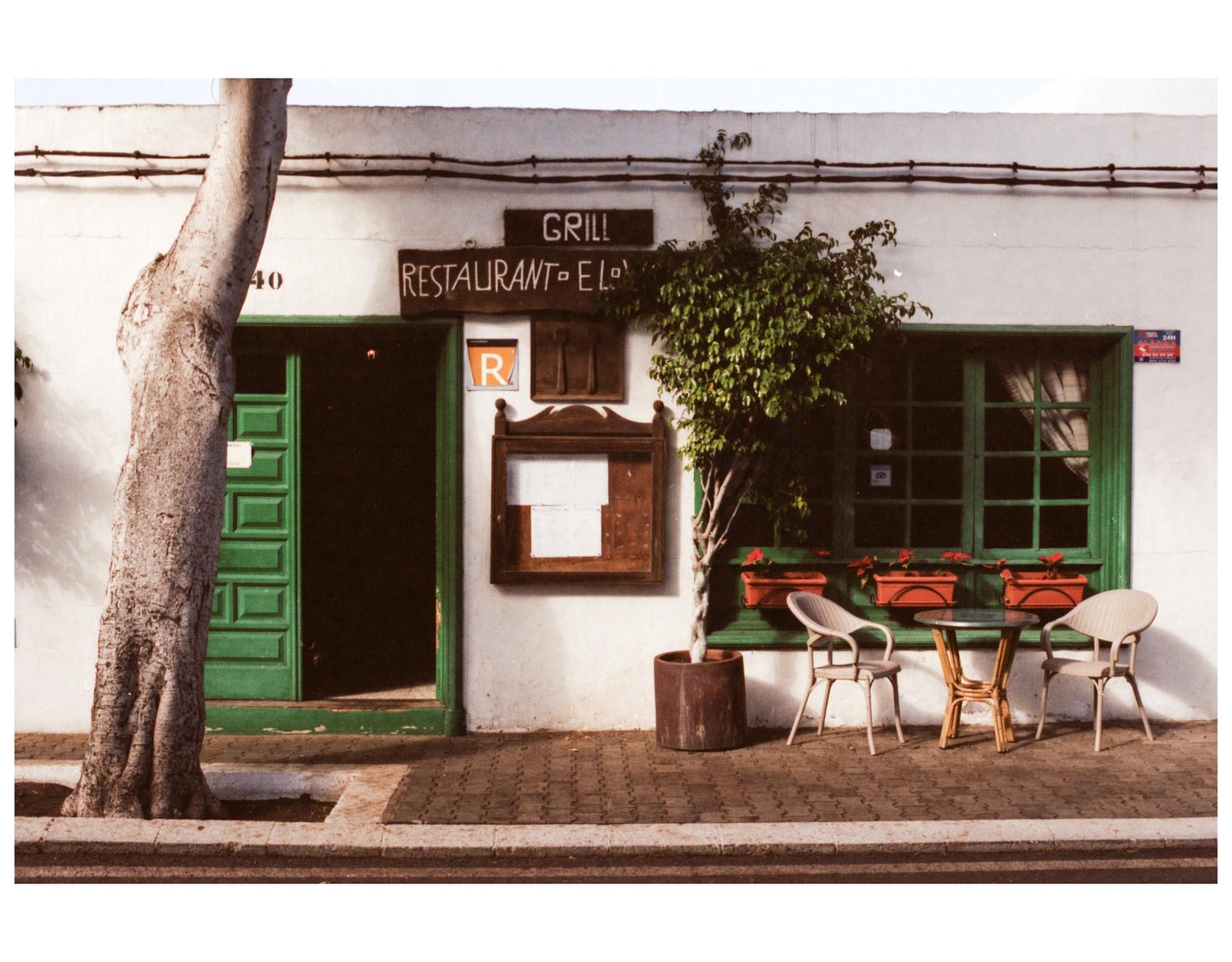 A tree stands in front of a white building with a green door and window, and a small table with two chairs sits on the sidewalk.