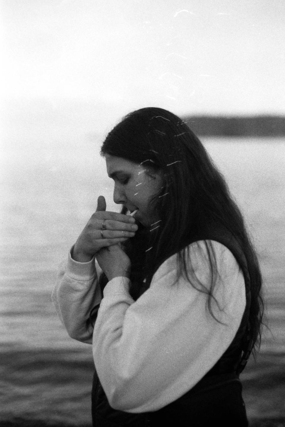 A woman with long hair lights a cigarette by the water.