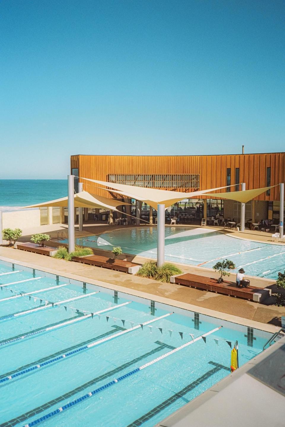 A swimming pool with lane lines is in the foreground, with a building and the ocean in the background.