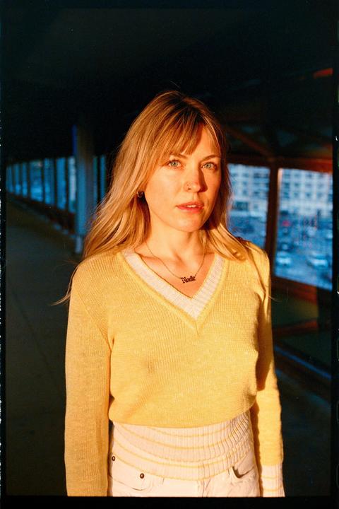 A woman wearing a yellow sweater and a necklace stands in a dark hallway with a window looking out onto a city street.