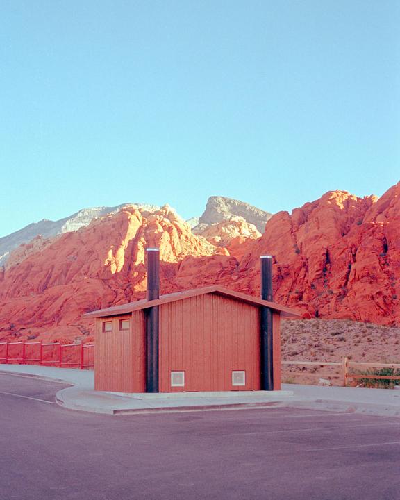 A small building with two tall black pipes stands on the side of a road with red rock mountains in the background.