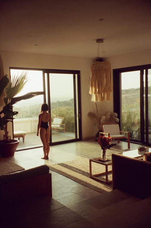 A woman stands in a sunlit room looking out through large glass doors to a landscape beyond.