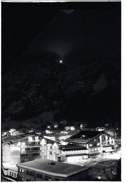 A small town with snow-covered roofs is nestled at the base of a dark, snow-dusted mountain at night.