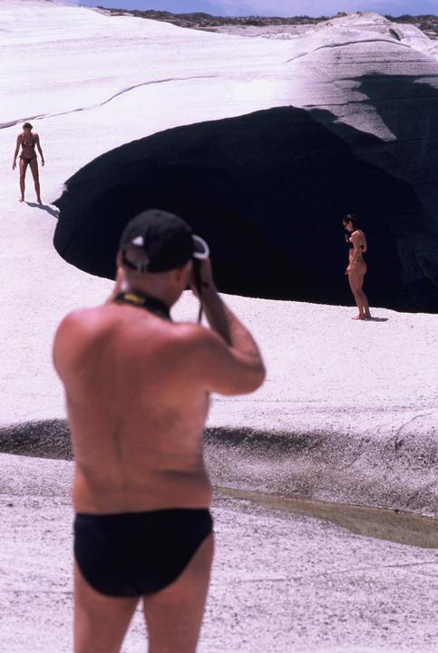 A man photographs two women in bikinis on a white rocky beach with a dark cave entrance.