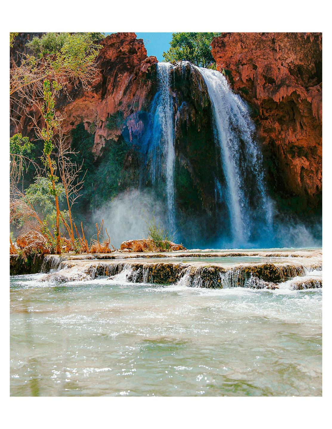 A large waterfall cascades over red rock formations into a series of pools and a river.