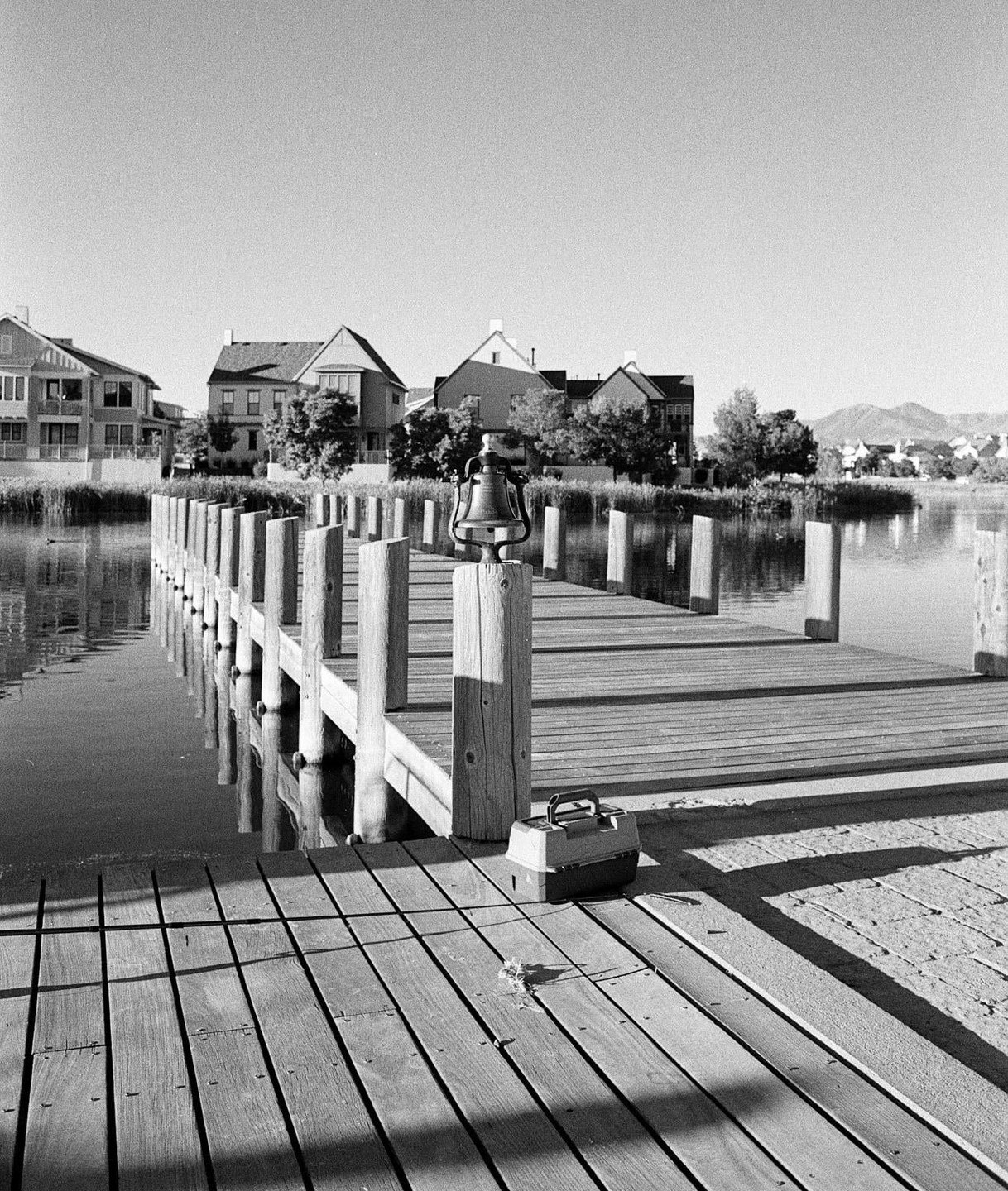 A wooden dock with a bell and a tackle box extends into a calm body of water, with houses and trees visible on the opposite shore under a clear sky.