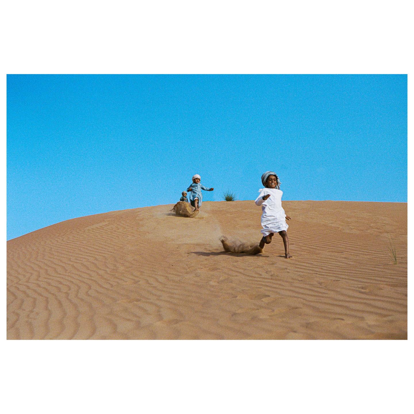 Three children play on a sand dune under a clear blue sky.
