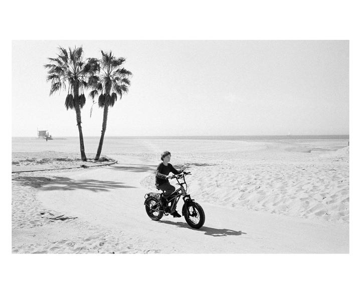 A boy rides a bicycle on a paved path next to a sandy beach with palm trees and the ocean in the background.
