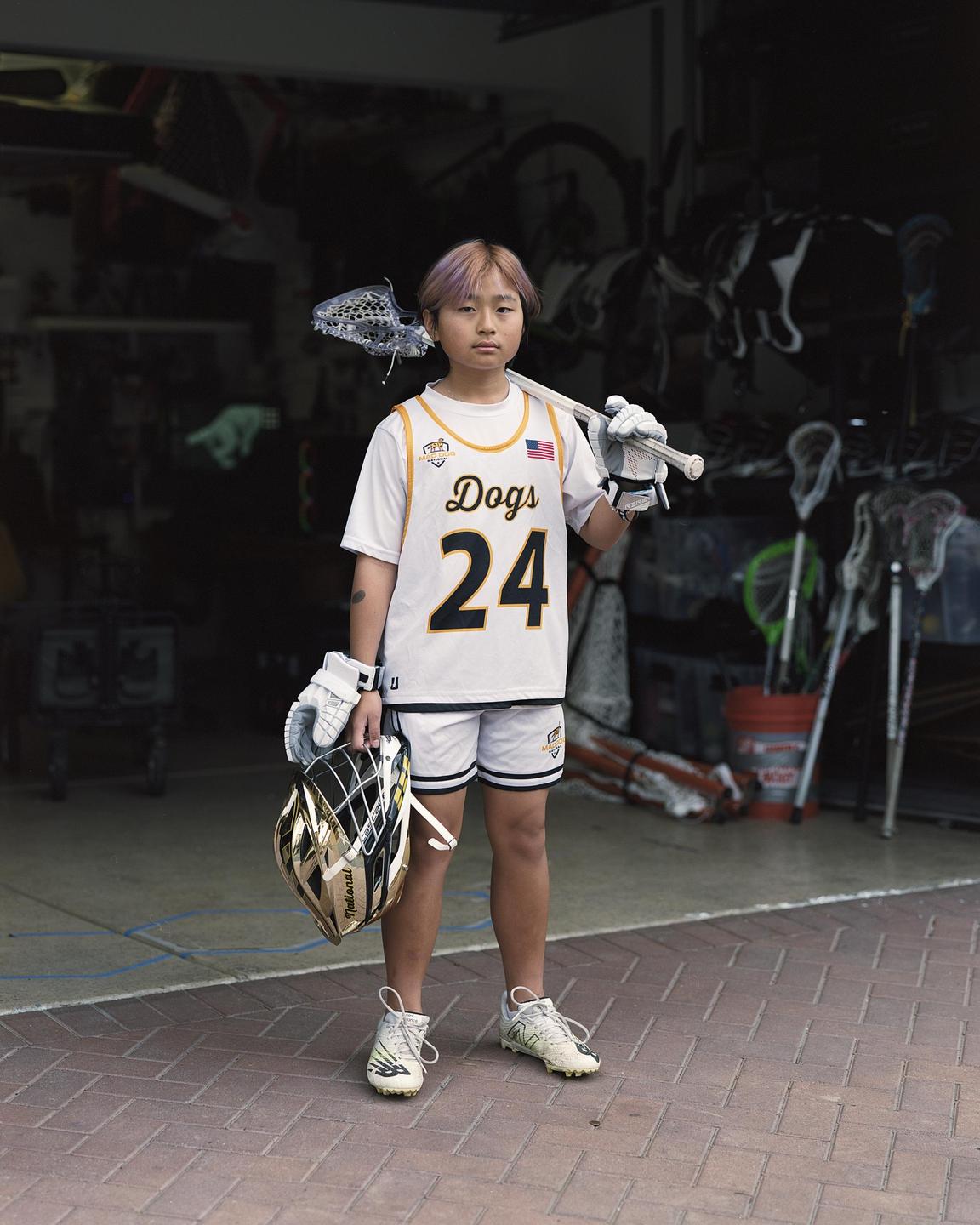 A young lacrosse player stands with their equipment in front of a garage.