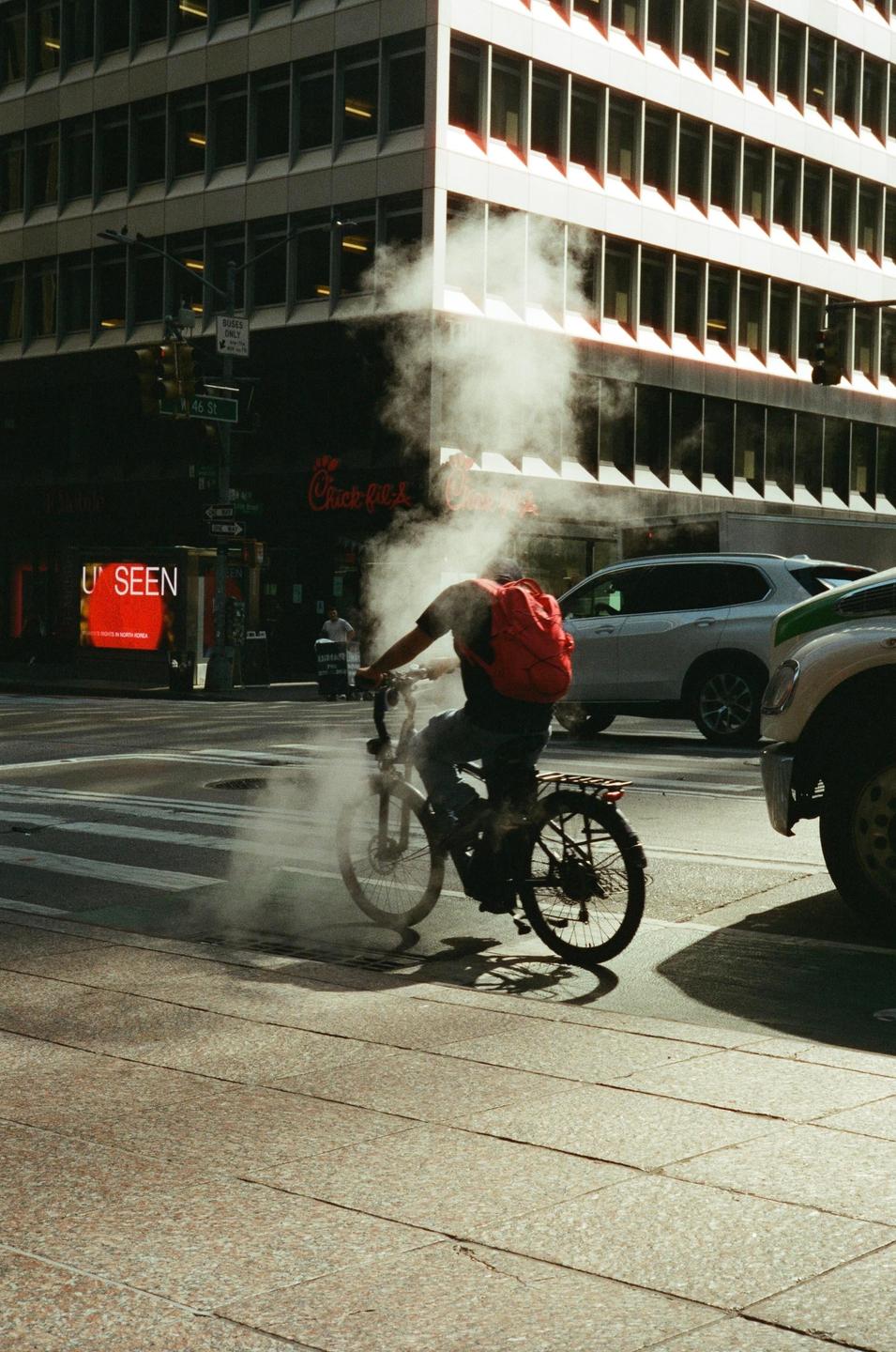 A man on a bicycle rides through a cloud of steam on a city street.