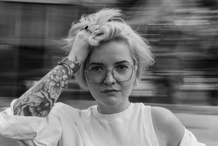 A woman with short blonde hair, glasses, and a tattooed arm looks directly at the viewer.