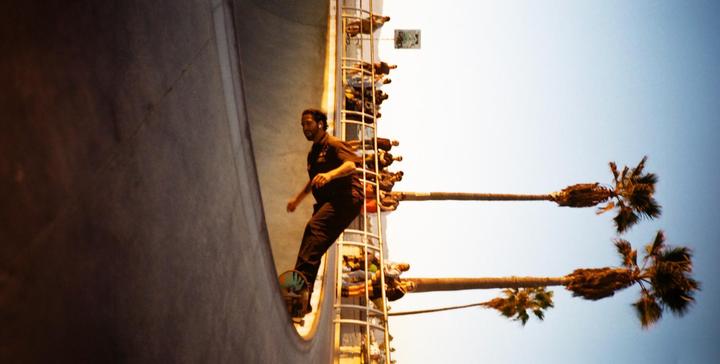A man on a skateboard is in a skate park with palm trees and spectators in the background.
