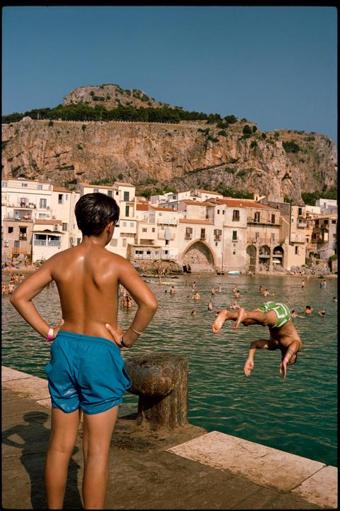 A boy in blue swim trunks stands on a pier watching another boy dive into the water in a bay surrounded by buildings and a large mountain.